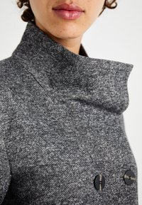 Grå frakke lavet af tekstureret stof; har en høj krave, dobbelte knaplukninger med sorte knapper og et struktureret design.