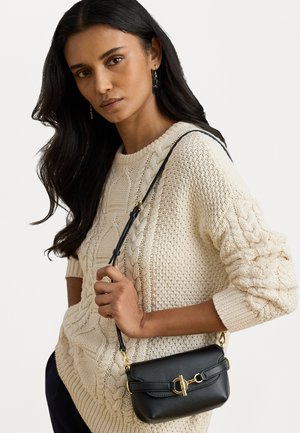 Černá kožená crossbody taška obdélníkového tvaru se zlatými detaily a štíhlým popruhem. Nosit se přetahuje přes krémový svetr s pleteným vzorem.