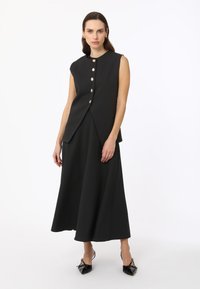 Zwart mouwloos jurk met een aansluitende taille, een uitlopende rok en grote gouden knopen. Matte textuur. Een paar zwarte schoenen versierd met een strik.