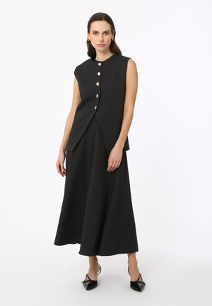 Black sleeveless dress with a fitted bodice, flared skirt, and large gold buttons. Matte texture. Pair of black bow-embellished shoes.