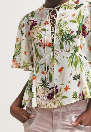 Blouse à motif floral avec laçage devant, manches courtes évasées et bas évasé, associée à un pantalon clair avec des poches avant décoratives.