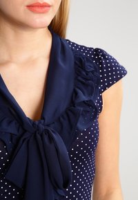 Mörkblå polka dot-blus med volangkrage och rosett vid halsringningen, med korta puffärmar och en mjuk tygkvalitet.