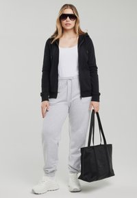 Kobieta w czarnej bluzie na zamek, białym topie, szarych spodniach dresowych, białych tenisówkach, z czarnymi dużymi okularami przeciwsłonecznymi, trzymająca czarną torbę na ramię.