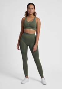 Olivgrön sport-bh och högmidjade leggings, tillverkade av texturerat tyg med kontrasterande accenter och meshpaneldetaljer. Vita sneakers.