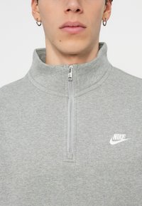 Šedá mikina na zip vyrobená z měkké tkaniny, která má vysoký límec, přední zip a malé bílé logo Nike na levé části hrudníku.