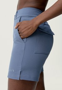 Close-up of a person wearing fitted blue shorts with a hand in the front pocket and a side flap pocket visible.