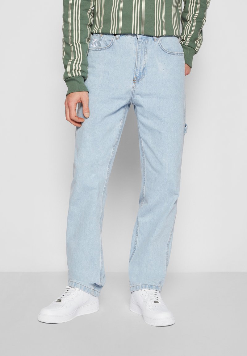 Vaqueros de mezclilla celestes con corte recto, cinco bolsillos y un leve desgaste, combinados con zapatillas blancas y una camisa de manga larga a rayas verdes.