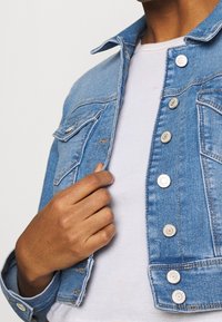 Ljusblå denimjacka med metallknappar, bröstfickor och en texturerad finish. Modell som håller jackan med en vit skjorta under.