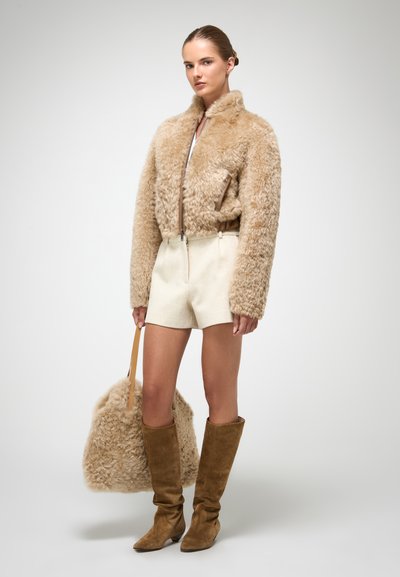 Brun faux-fur jakke med høj krave, cremede shorts, brune ruskinds knæhøje støvler og en matchende fluffy taske. Glatte teksturer dominerer.