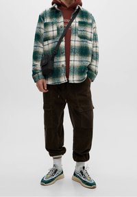 Camicia in flanella a scacchi verde e bianca sopra una felpa marrone, abbinata a pantaloni in corduroy marroni e scarpe da ginnastica multicolori. Piccola borsa a tracolla nera.