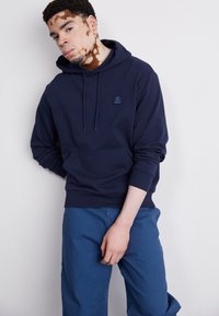 Granatowy hoodie z kieszenią z przodu i kapturem na sznurki, z małą łatą z logo. W połączeniu z niebieskimi spodniami, które mają luźny krój.