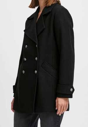 Kobieta w czarnym dwurzędowym płaszczu typu pea coat z srebrnymi guzikami, bocznymi kieszeniami i zapinanymi mankietami, w połączeniu z ciemnymi dżinsami.