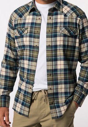 Mann trägt ein blau-grün-creme kariertes Hemd mit Knöpfen über einem weißen T-Shirt und beige Hose mit Kordelzug, Hände nahe den Taschen.