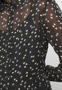 Svart genomskinlig blus med gula och vita hjärtformade mönster, med knäppning fram, krage och rynkade detaljer vid midjan.