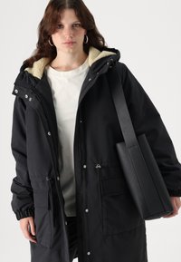 Parka noire avec un intérieur en beige peluche. Dispose d'une fermeture à glissière et de pressions, de poignets élastiques et de deux poches avant. Sac noir à la main.