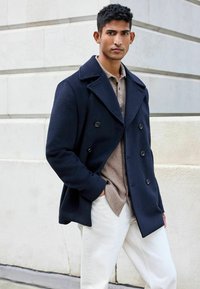 Trinchera de lana azul marino con botones de doble pecho, solapas anchas y bolsillos laterales, combinada con una camisa clara de botón y pantalones blancos.