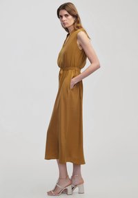 Senf-farbenes ärmelloses Jumpsuit mit V-Ausschnitt, taillierten Bund, Seitentaschen und fließendem Stoff; kombiniert mit klobigen weißen Absatzsandalen.