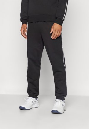 Mann trägt schwarze Jogginghose mit weißen Seitenstreifen und weißen Sportschuhen, steht vor einem schlichten hellen Hintergrund.