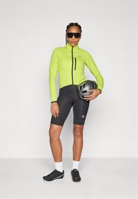 Strój rowerowy z dopasowaną, fluorescencyjnie zieloną kurtką z zamkiem błyskawicznym oraz czarnymi spodniami z podszewką. Dodatki to szary kask i okulary przeciwsłoneczne.