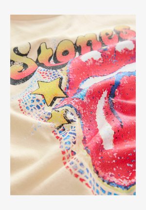 Pale sárga pamut póló, amelyen piros ajkak, csillagok és absztrakt minták kék és fekete színben találhatók, texturált hatással.