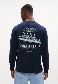 Tumesinise pika varrukaga särk, mille seljal on nelja aerutaja graafiline trükk ning tekst "Tõeline Stockholmi Sportlaste Klubi."