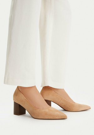 Beżowe zamszowe buty na słupku z noskiem w szpic, noszone z białymi szerokimi spodniami na neutralnym tle.