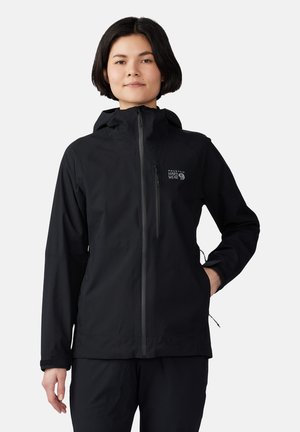 Person trägt eine schwarze Mountain Hardwear Kapuzenjacke mit Reißverschlusstasche auf der Brust und Händen in den Seitentaschen vor weißem Hintergrund.