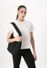 Zwarte gewatteerde schoudertas met een brede strap, gedragen over een wit t-shirt met een klein rood hart. Model draagt zwarte leggings.
