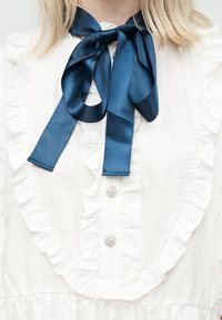 Hvit bluse med rysjekant, krystallknapper og en knyttet navyblå satengsløyfe ved kragen.