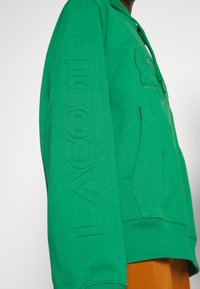 Sudadera verde hecha de una mezcla de algodón, con un gran logo de cocodrilo bordado, bolsillo lateral y "LACOSTE" grabado en la manga.