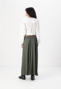 Blouse blanche à manches longues avec un détail froncé dans le dos, associée à une longue jupe maxi fluide de couleur vert foncé. Bottines noires à boucles.