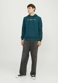 Sudadera verde azulado con cordones y bolsillo frontal, que presenta el logo "JACK & JONES" en naranja. Combinada con unos vaqueros grises de corte holgado y zapatillas negras.