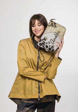 Frau in senffarbener Jacke hält eine Tiermuster-Tasche mit der Aufschrift "Anekke" dicht an ihr Gesicht, lächelt mit geschlossenen Augen.