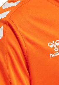 Tissu orange vif avec logo abeille Hummel blanc et bandes en chevron sur la zone des épaules d'un maillot ou d'un t-shirt de sport.