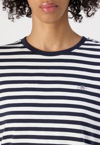 Personne portant une chemise à rayures horizontales bleu marine et blanches avec un petit logo brodé sur la poitrine gauche et des cheveux bouclés visibles sur les bords.