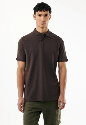 Junger Mann, der ein dunkelbraunes Poloshirt mit kurzen Ärmeln und olivgrüne Shorts trägt, steht vor einem schlichten weißen Hintergrund.