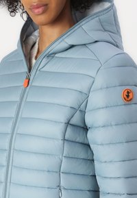 Lichtblauwe pufferjack met horizontaal gestikt ontwerp, een capuchon, een ritssluiting aan de voorkant en een opvallende oranje logo patch op de mouw.