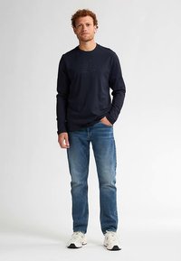 Marineblauw shirt met lange mouwen en ronde hals, voorzien van een subtiel grafisch detail, gecombineerd met lichtblauwe spijkerbroek en witte sneakers.