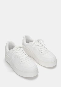 Białe sneakersy z gładką skórzaną cholewką, okrągłym noskiem, perforowanym wzorem, płaskimi sznurowadłami i grubą gumową podeszwą dla dodatkowej wysokości.