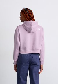 Haut de hoodie coupé en lavande clair avec une capuche, des poignets côtelés et un petit logo blanc dans le dos. Associé à un pantalon bleu marine.