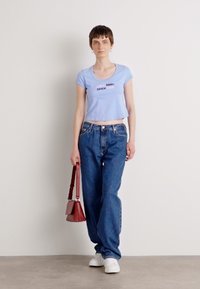 Light blue cropped T-shirt with "Calvin Klein since 1980" text, paired with loose blue jeans and white sneakers. Holding a red handbag.
