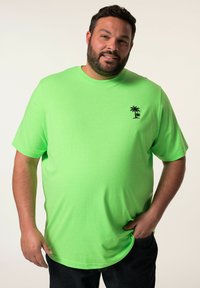 Svěží zelené tričko s krátkým rukávem, vyrobené z měkkého materiálu, s malým černým grafickým motivem palmy na levé straně hrudníku, v kombinaci s tmavými džíny.