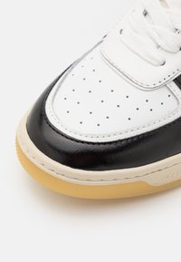 Närbild av en svart och vit sneakers tå med perforerat vitt läder, svarta detaljer, vita snörningar och en ljusbrun gummisula.