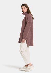 Fleece pullover i mauve med høj krave, lange ærmer og sideslids for bedre bevægelighed. Parret med cremede bukser og lyse beige sneakers.