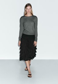 Pull gris à manches longues et jupe noire à volants superposés. L'ensemble met en avant une taille cintrée et une texture fluide, associé à des sandales noires.