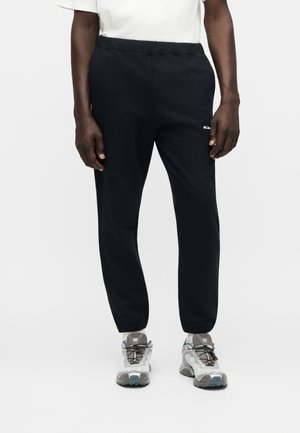 Homme portant un jogging noir à taille élastique et des baskets blanches, debout devant un fond clair uni.