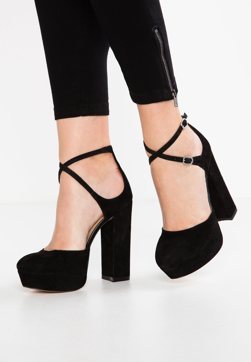 Černé semišové lodičky na vysokém podpatku s robustním platformou, vybavené křížovými řemínky kolem kotníku a štíhlým vertikálním designem podpatku.
