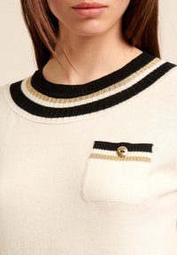 Maglione color crema con colletto a righe nere, bianche e oro. Presenta una piccola tasca con un bottone e un bordo nero di contrasto.