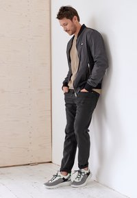 Veste bomber noire à fermeture éclair avec des poignets côtelés, pull kaki, pantalon noir slim et baskets grises avec accents verts. Design minimaliste.