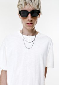Person med kort blond hår iført store sorte solbriller, en hvid skjorte med knapper og to sølvkæder mod en hvid baggrund.